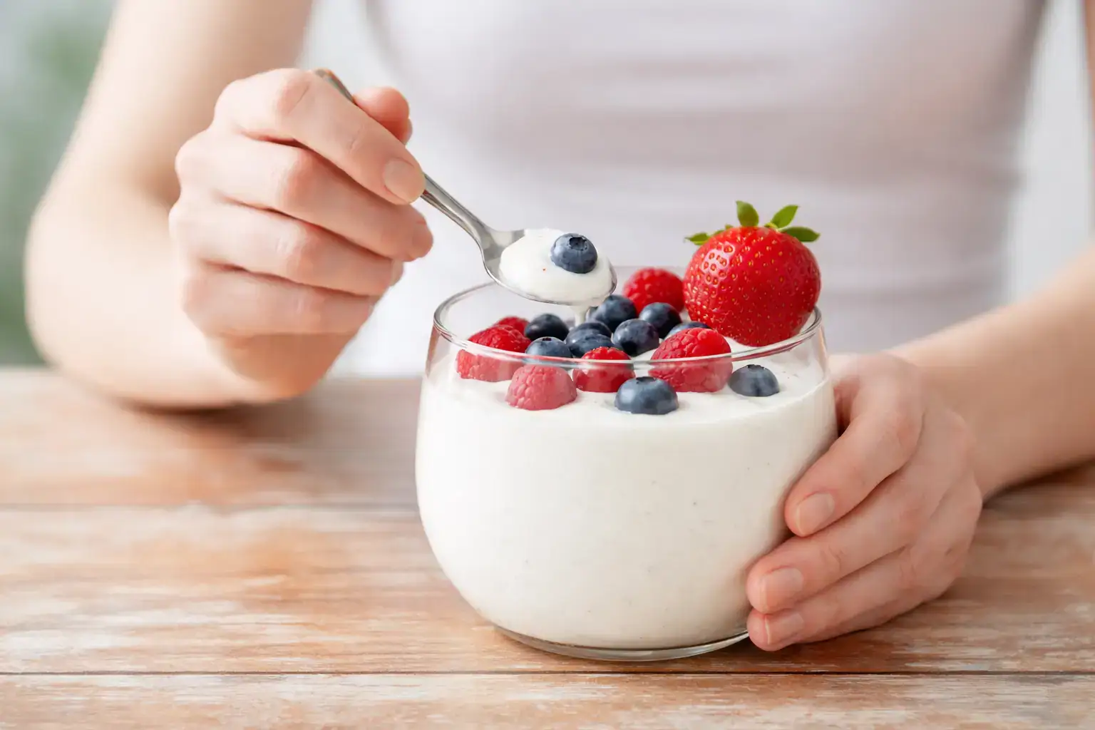 Nahaufnahme einer Frau, die ein Glas mit Joghurt und frischen Beeren (Erdbeeren, Himbeeren, Blaubeeren) hält. Sie löffelt eine Portion mit einem silbernen Löffel.